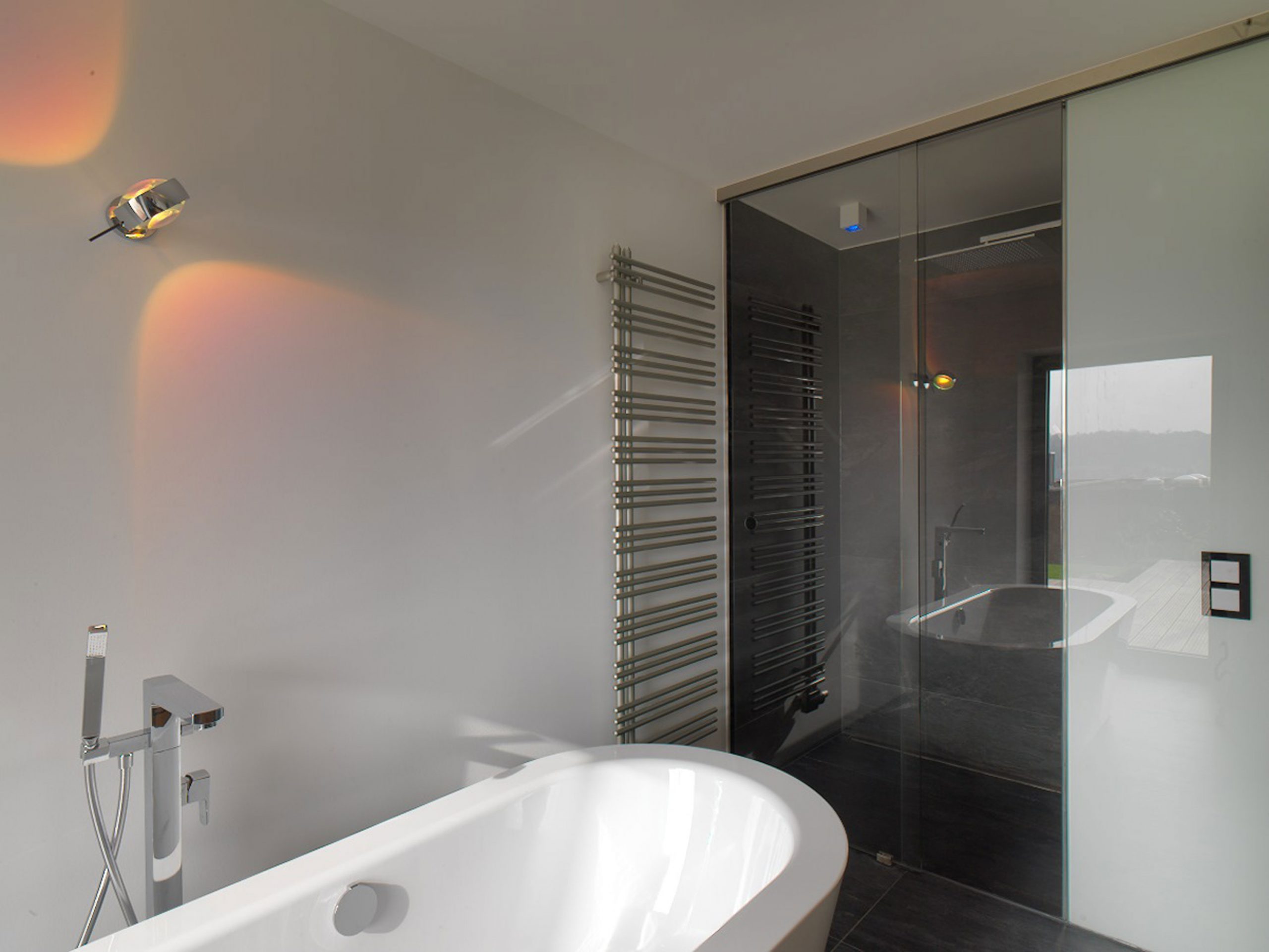 Die freistehende Badewanne im Haus KMH mit Blick auf den durch Glas-Schiebeelemente abgetrennten Duschbereich mit Duschhimmel.